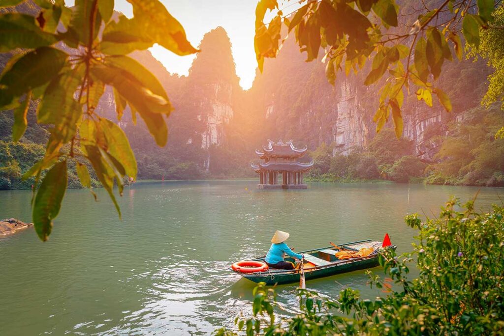 Small boat passing a temple in Trang An, Ninh Binh, surrounded by limestone cliffs and lush greenery in soft morning light