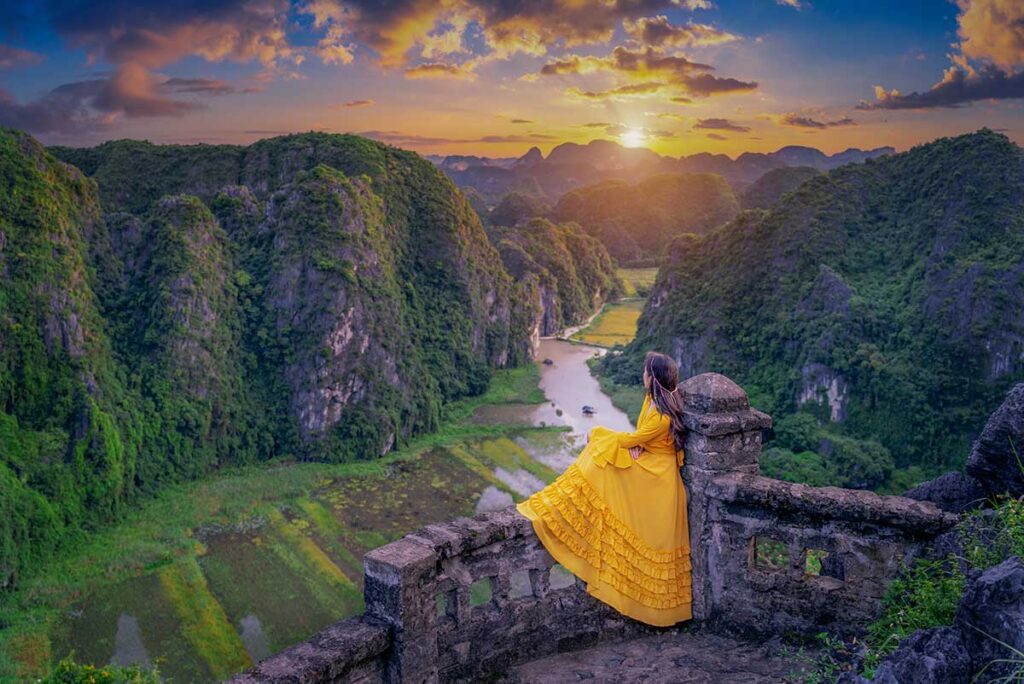 Woman in a yellow dress overlooking the Tam Coc valley from Mua Cave viewpoint in Ninh Binh at sunset, with limestone karsts and rice fields below