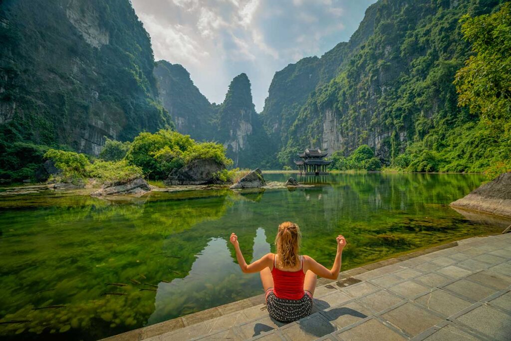 Traveler sitting by a calm lake in Trang An, Ninh Binh, surrounded by limestone cliffs and greenery with a small temple in the distance