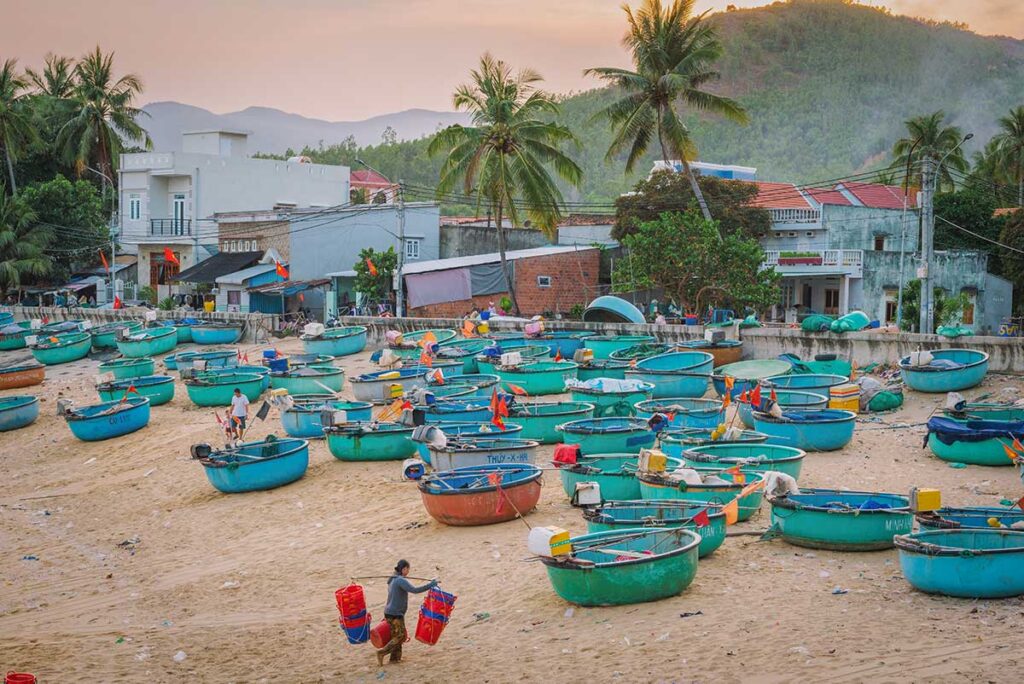 Phu Yen fishing village with dozens of round basket boats on the sandy beach, palm trees, and houses set against green hills at sunset.