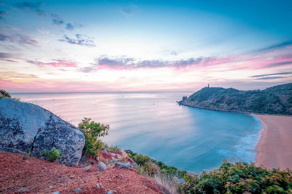 Curved beach and coastline in Phu Yen seen from above, with soft pastel sky and calm sea at sunrise