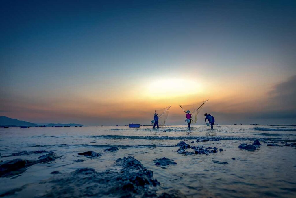 Local fishermen casting nets in shallow water at sunrise in Phu Yen, with soft pastel sky and calm sea creating a peaceful coastal scene