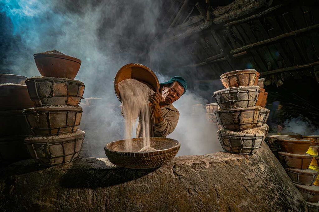 Local artisan in a Phu Yen salt-making village pouring fresh salt into bamboo baskets inside a smoky workshop.