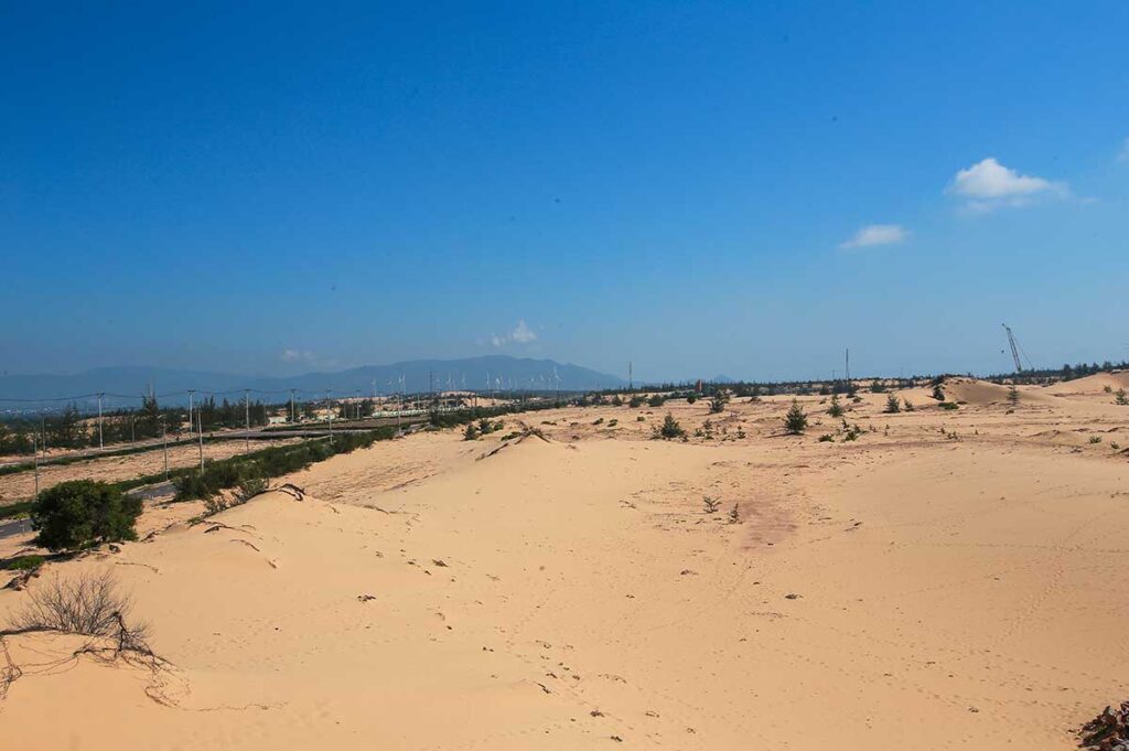 Expansive desert-like scenery of Phuong Mai Sand Dunes in Binh Dinh Province with a view towards the mountains and rows of wind turbines on the horizon