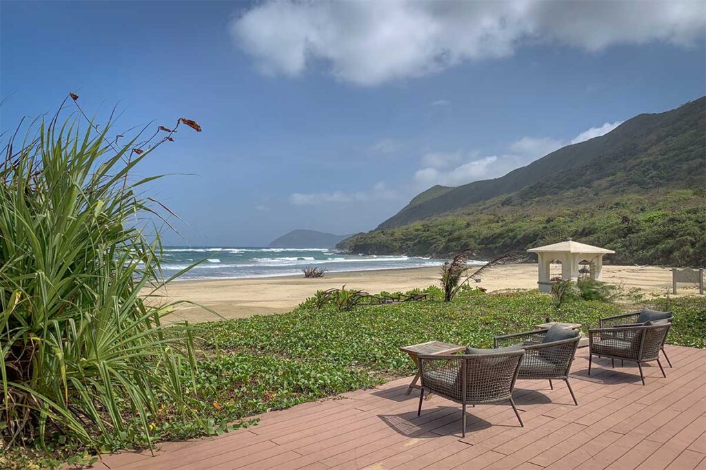 Dong Beach in Con Dao as seen from Poulo Condor Resort, with lounge chairs on a terrace overlooking a wide sandy beach and waves along the coastline.