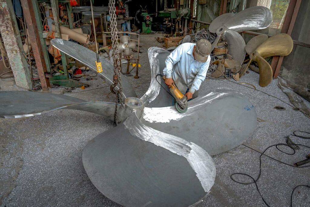 Skilled Vietnamese craftsman polishing a large ship propeller in a workshop in Rach Gia, part of the local boat-building industry in the Mekong Delta.