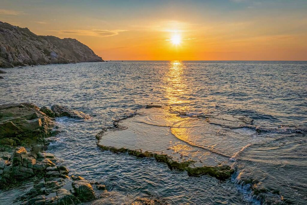 Rai Cave in Ninh Thuan at sunrise, waves crashing over flat rock formations along the dramatic coastline.”
