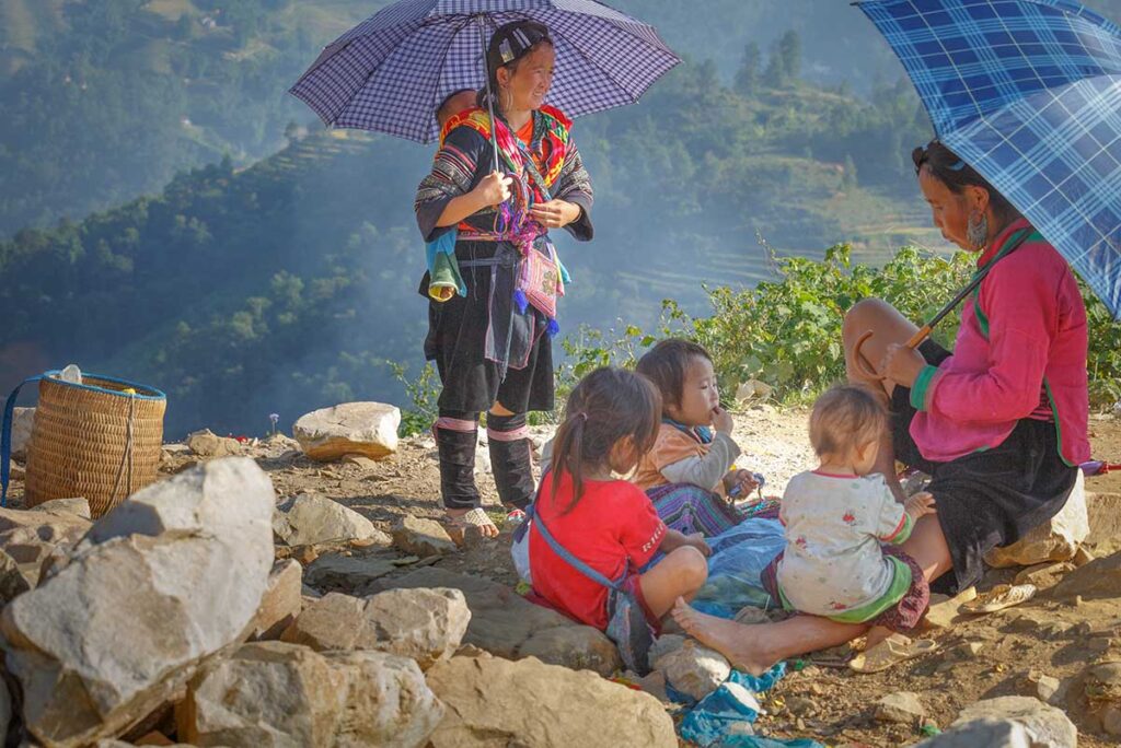 Ethnic minority women and children in Sapa resting on a hillside, wearing traditional clothing and holding umbrellas