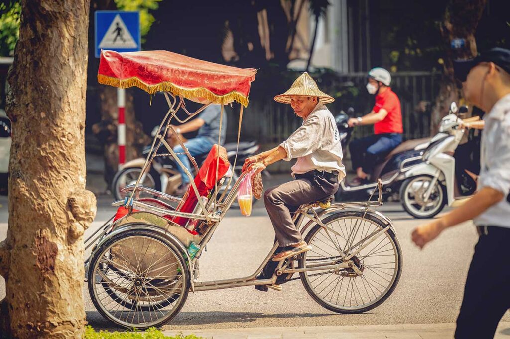 Man riding a cyclo through a busy street in Vietnam, with motorbikes and pedestrians passing in the background