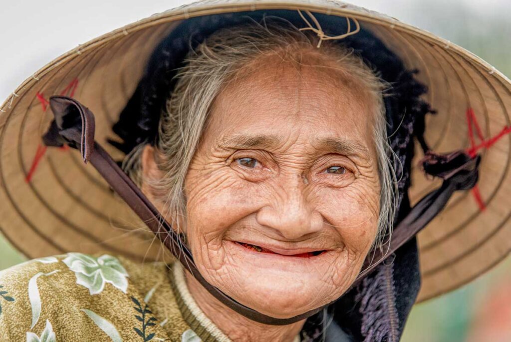 Close-up portrait of an elderly Vietnamese woman wearing a conical hat, showing detailed facial features and a warm smile