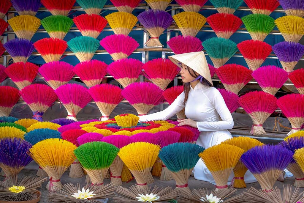 Woman in a white ao dai arranging colorful incense sticks in a traditional village setting, creating a vibrant and symmetrical scene