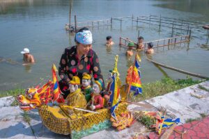A woman sitting on the edge of the water with water puppets and boat prop with local man setting up a water puppet theater in the water behind her at Water Puppet Village in Thanh Hai Commune