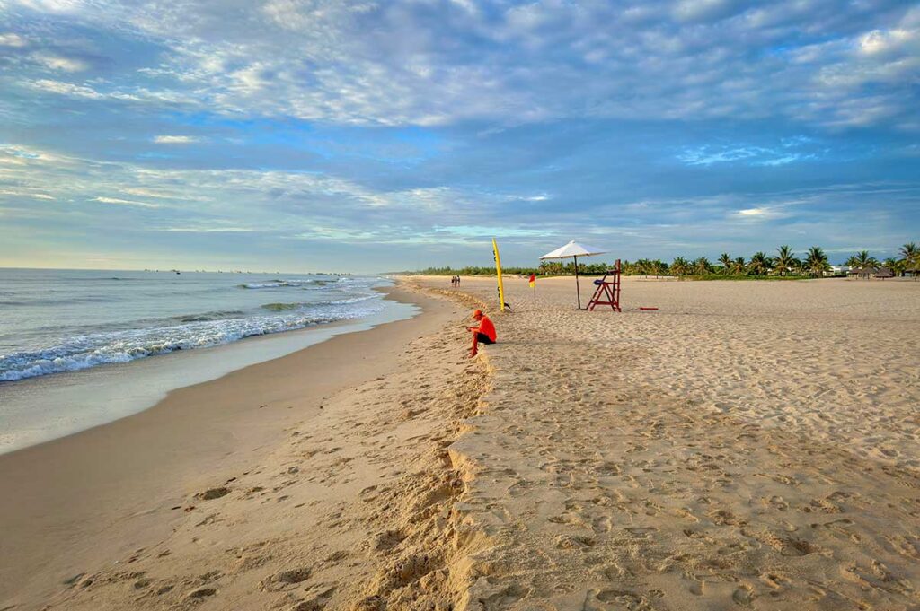 Calm sea and long sandy coastline at Binh Minh Beach in Quang Nam, with resorts and palm trees in the background.