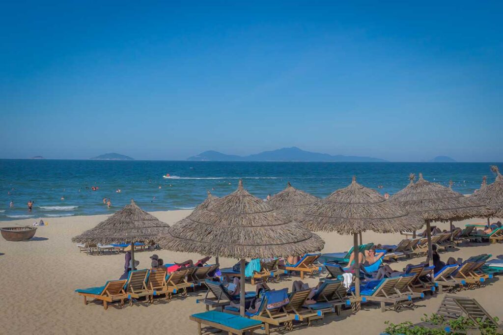 Travelers relaxing at Hoi An’s An Bang Beach in August under clear blue skies, as warm sea temperatures and bright sunshine continue despite rising humidity.