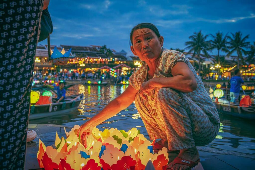 Local woman selling floating lanterns on the Thu Bon River in Hoi An during December, as night lights and reflections create a festive, peaceful scene.