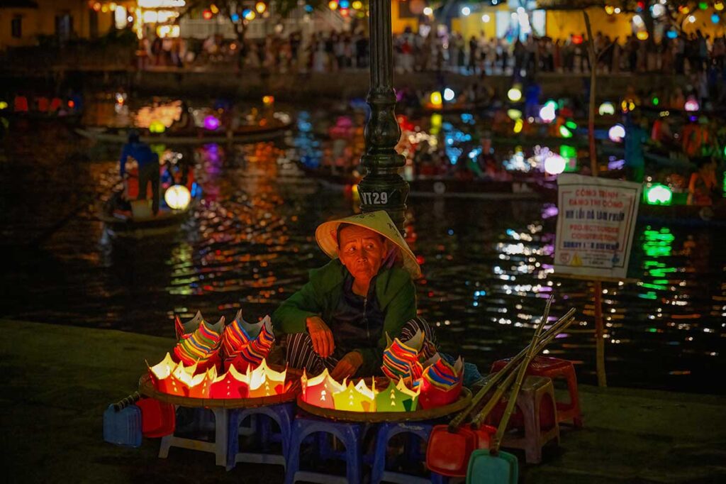 Elderly vendor selling paper lanterns by the river during the Hoi An Lantern Festival, with colorful lights reflecting on the water and boats in the background.