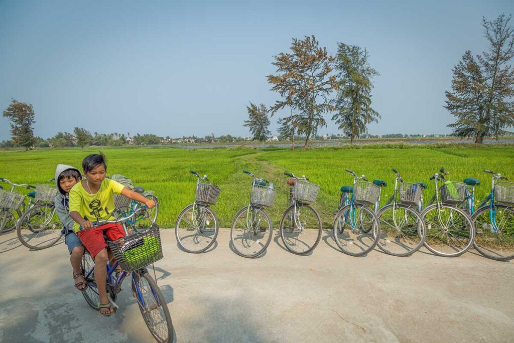Local children cycling through green rice fields near Hoi An in March, when the weather turns warm and dry—ideal for countryside bike tours and exploring village life.