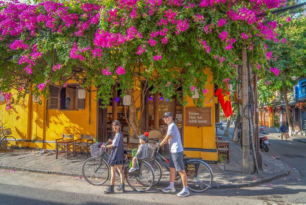 Family cycling through Hoi An’s old streets covered in blooming bougainvillea in May, when sunny days and colorful flowers mark the start of summer in central Vietnam.
