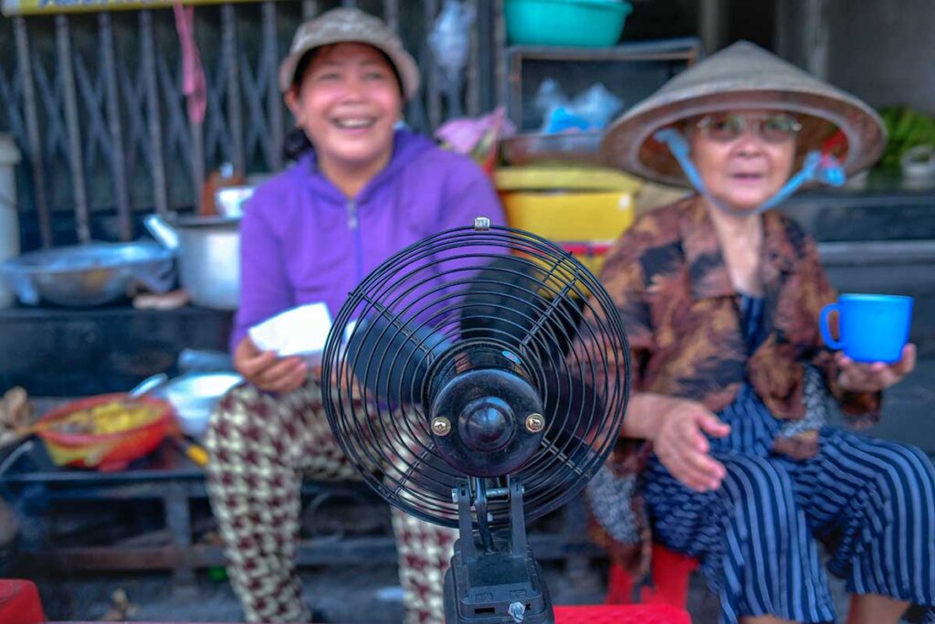 Two local woman sitting on the side of the street smiling with a small fan pointing towards them on a hot day in Hue
