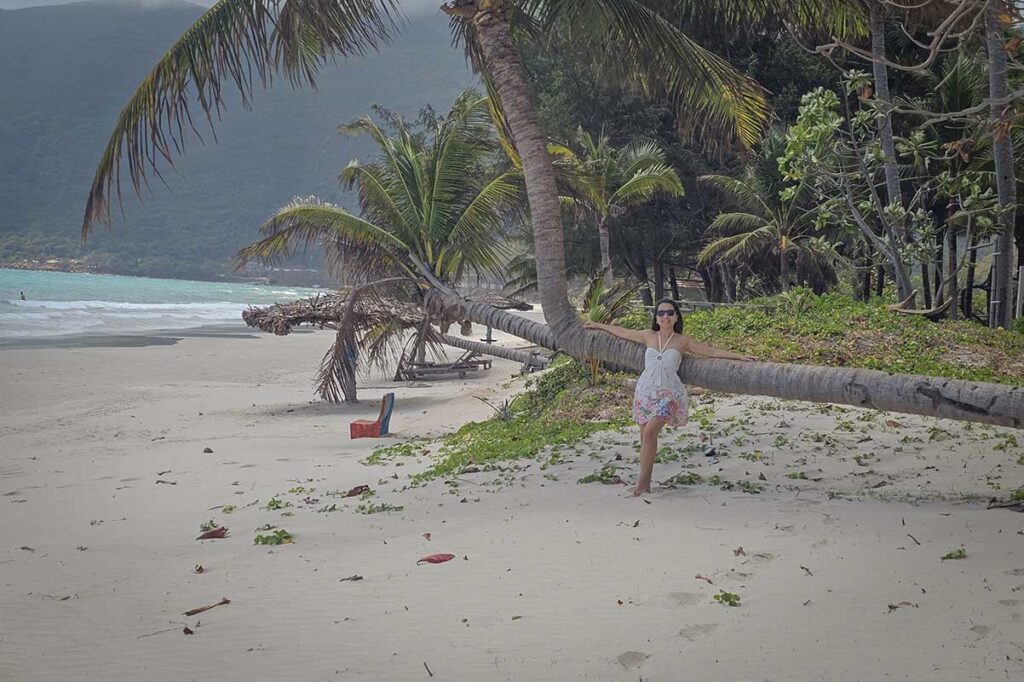 Woman standing beside a leaning palm tree at An Hai Beach in Con Dao with white sand, tropical vegetation, and calm sea behind.