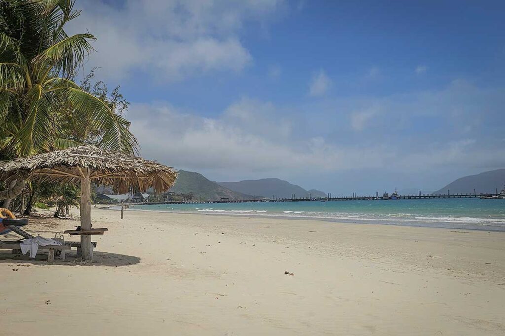 Palm trees and thatched beach umbrellas at An Hai Beach in Con Dao with calm turquoise water and a pier along the coastline.