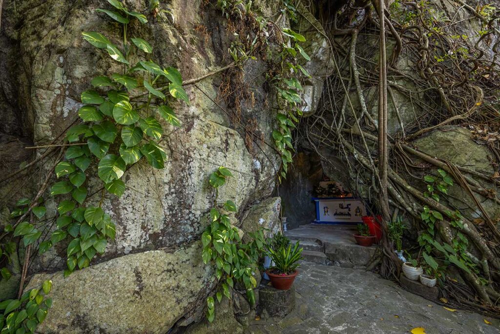 Small shrine built into a rocky cliff at Maria Cave in Con Dao National Park, surrounded by tree roots, vines, and lush jungle vegetation