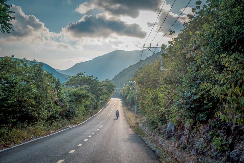 Quiet coastal road running through Con Dao National Park, with a motorbike driving toward forested mountains under a partly cloudy sky