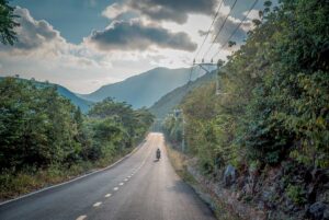 Quiet coastal road running through Con Dao National Park, with a motorbike driving toward forested mountains under a partly cloudy sky