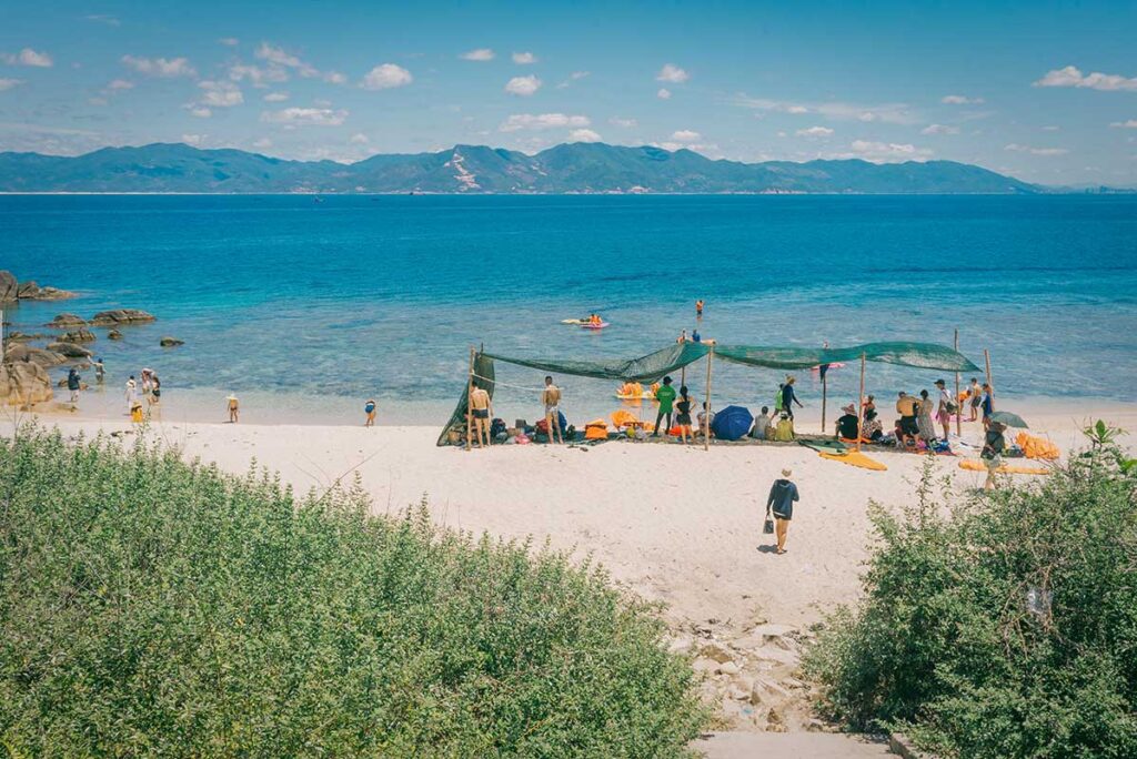 Small sandy beach on Cu Lao Xanh with visitors swimming and shaded nets for sun protection.