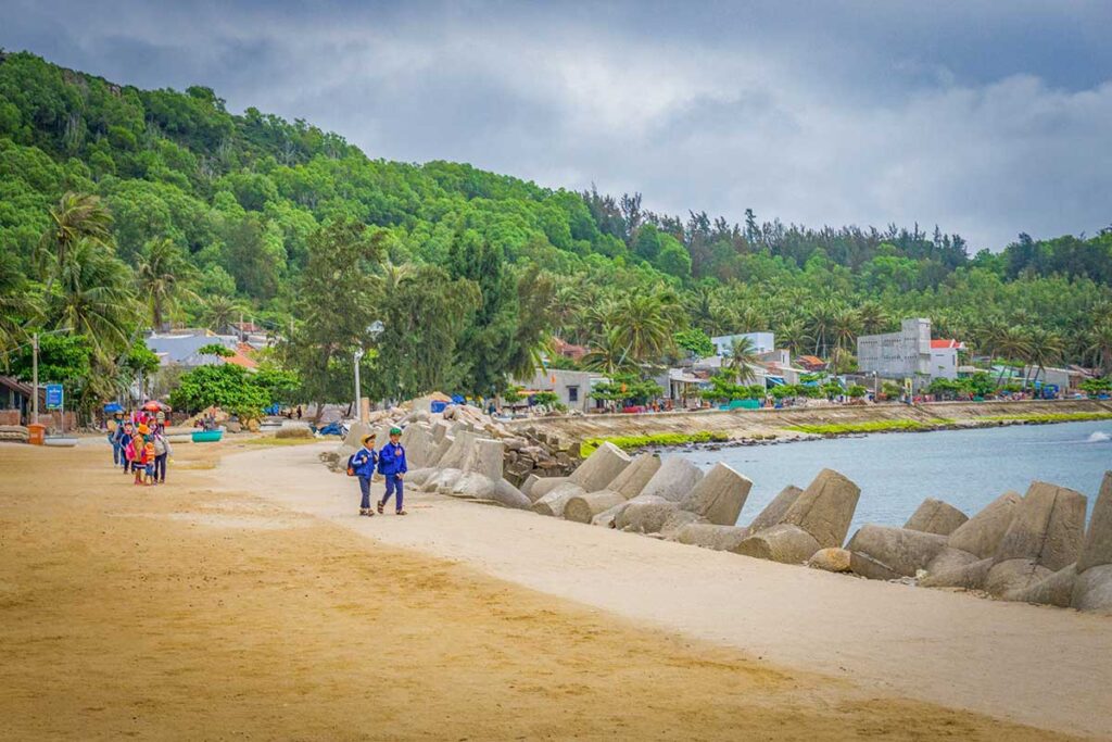 Children walking along the coast road of Cu Lao Xanh Island on their way to school, with palm trees and village houses behind.