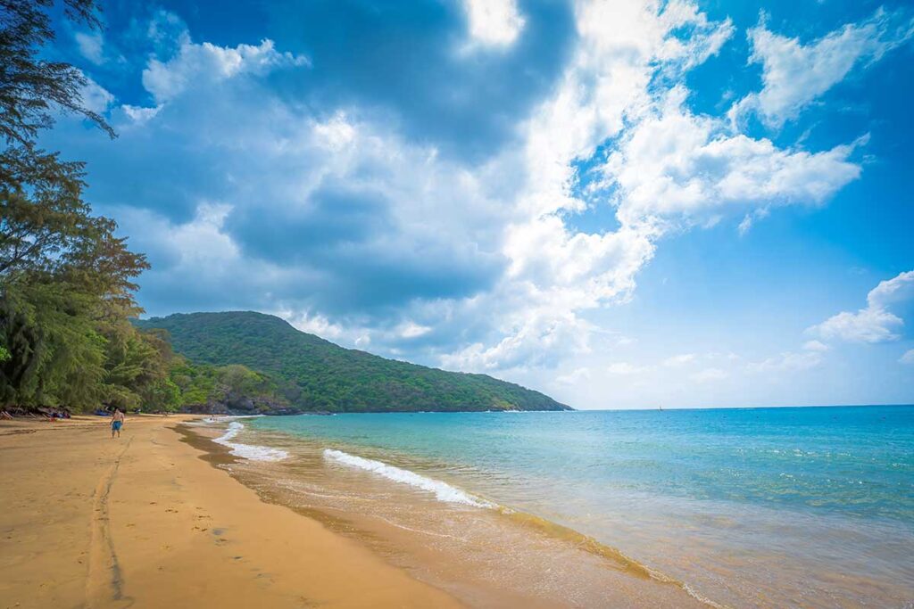 Wide sandy shoreline at Dam Trau Beach in Con Dao with turquoise water, gentle waves, and green hills under a dramatic cloudy sky.