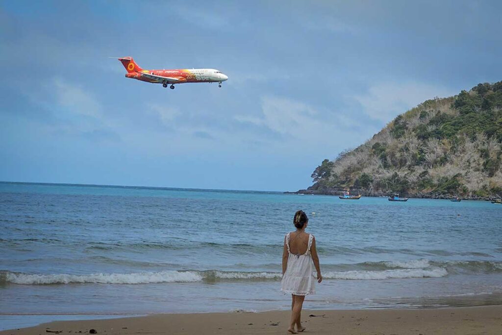 Woman walking along Dam Trau Beach in Con Dao while a plane flies low overhead during landing, with calm sea and boats in the background.