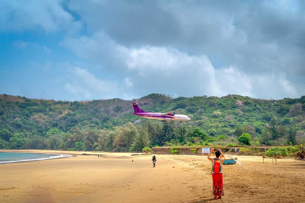 Low-flying passenger plane landing over Dam Trau Beach in Con Dao, with wide sandy shoreline and green hills in the background