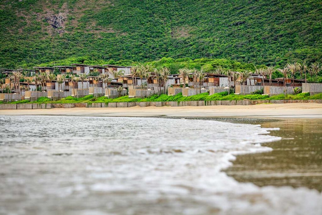 Seaside villas of Six Senses Con Dao overlooking Dat Doc Beach, with waves reaching the shore and lush hills rising behind the resort.