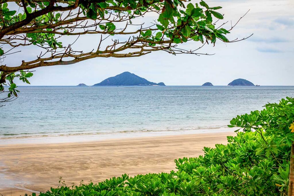 View of Six Senses Con Dao villas along Dat Doc Beach with palm trees, sandy shore, and luxury beachfront resort backed by green mountains.