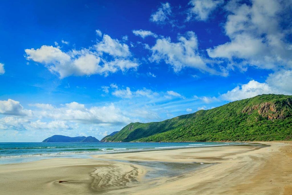 Dong Beach in Con Dao during low tide with wide sand patterns, shallow water, and surrounding mountains under a bright blue sky.