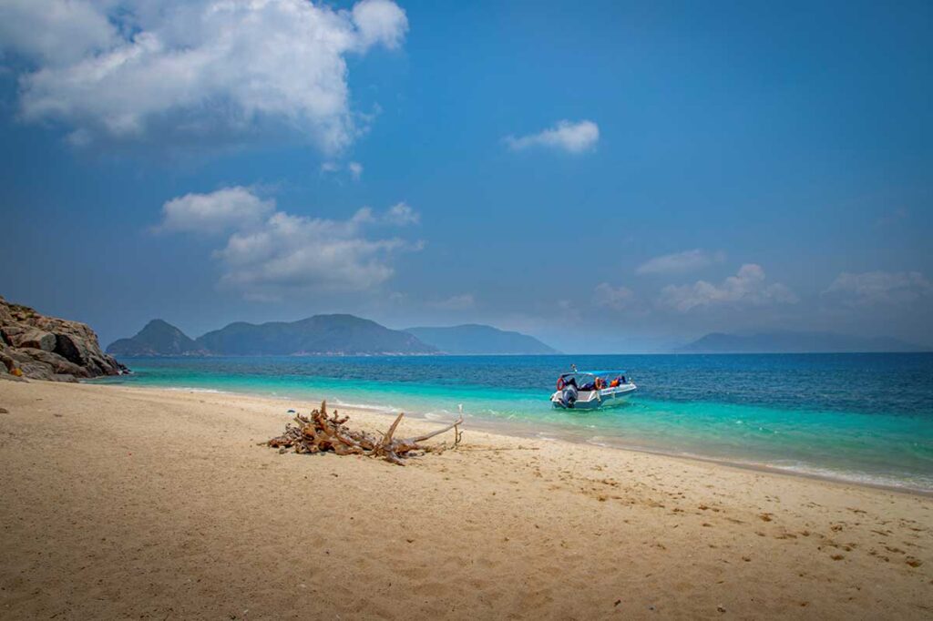 Quiet beach on Hon Cau Island in Con Dao with turquoise water, a small boat anchored near the shore, and mountains in the distance.