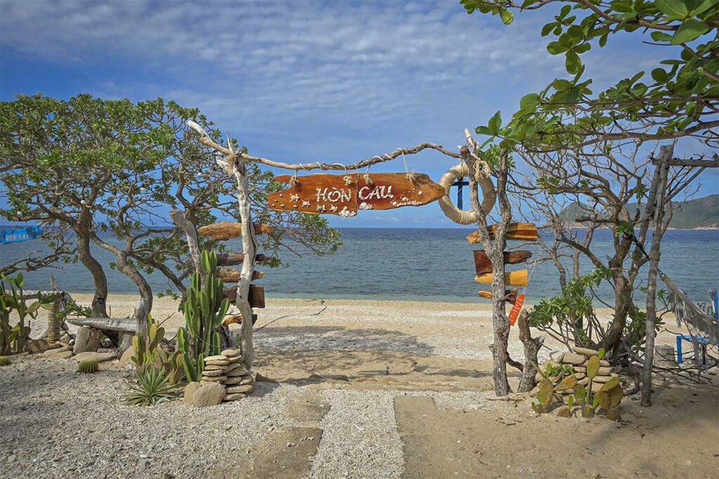 Entrance to Hon Cau Island beach in Con Dao with a rustic wooden sign, tropical plants, and the ocean visible in the background.