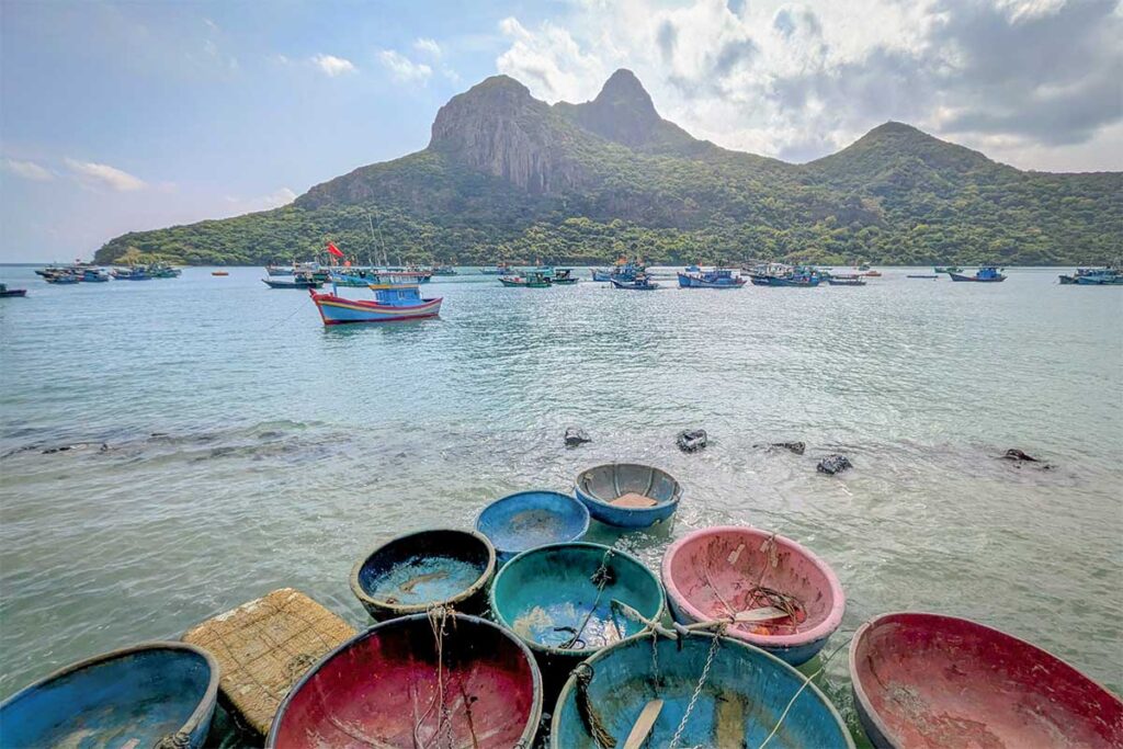 Fishing boats anchored near Hong Dam Island in Con Dao with calm sea, mountain backdrop, and traditional Vietnamese basket boats in the foreground.