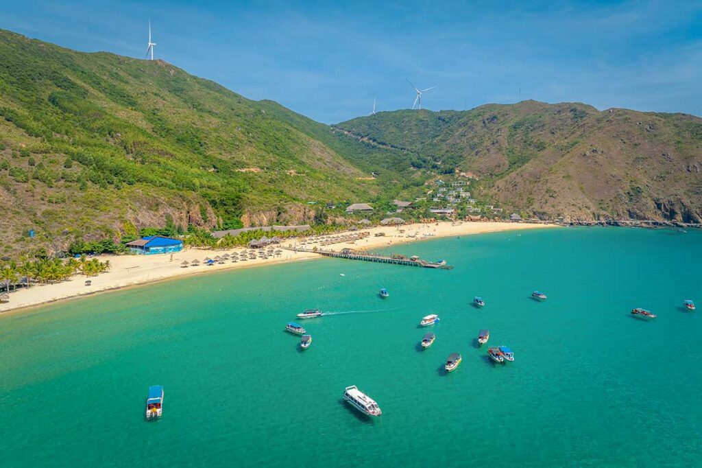 Aerial view of Ky Co Beach – Panoramic shot of the golden sand, clear turquoise water, and tour boats anchored along the cove.