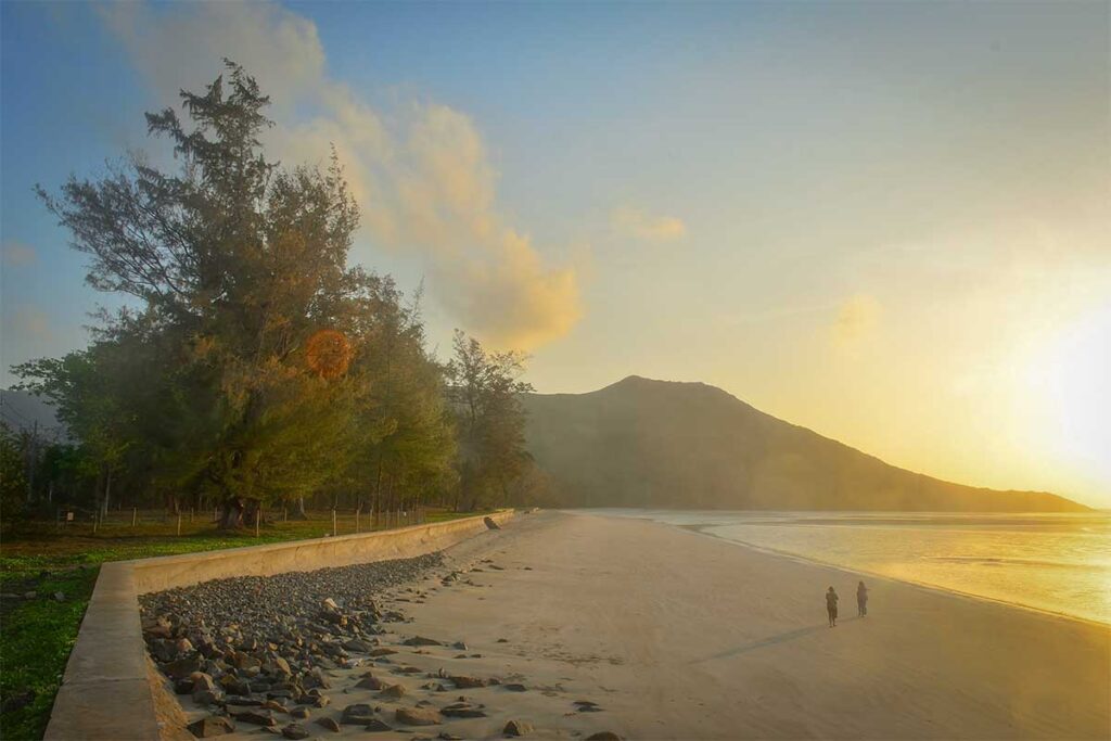 Sunset view of Lo Voi Beach in Con Dao with soft golden light, a wide sandy shoreline, and two people walking along the calm sea.