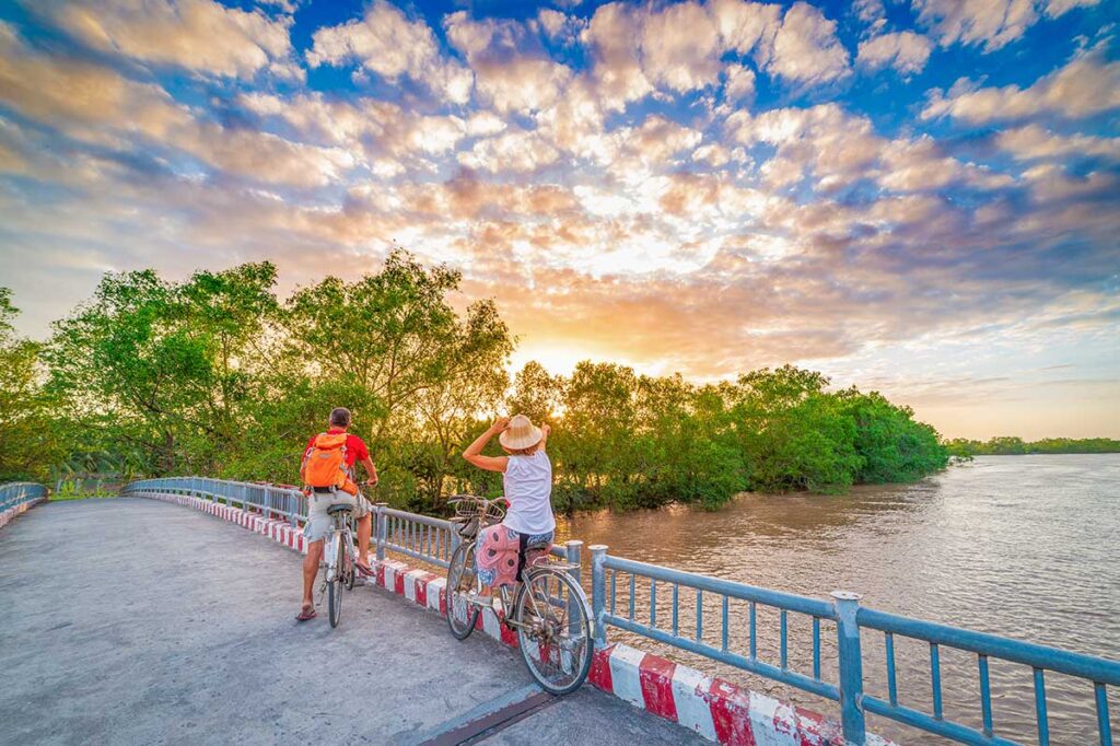 Couple cycling on a riverside bridge at sunset with golden clouds, capturing a beautiful cycling experience in the Mekong Delta.