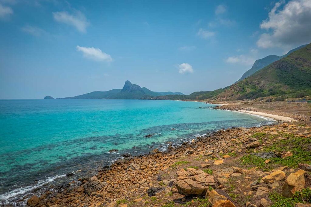 Panoramic view of Nhat Beach in Con Dao with clear turquoise water, rocky shoreline, and green hills surrounding the bay.