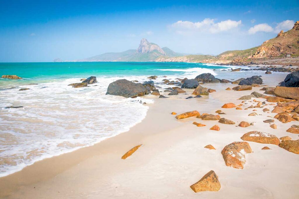Waves rolling onto Nhat Beach in Con Dao with golden sand, scattered rocks, and a mountainous coastline in the distance.