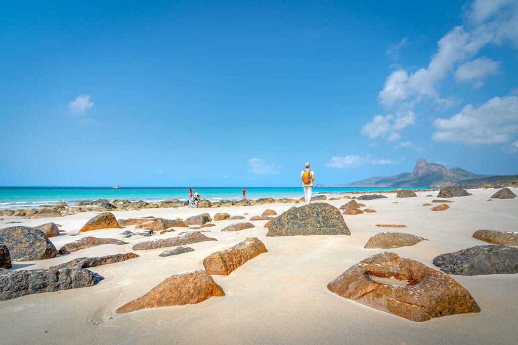 Wide sandy stretch of Nhat Beach in Con Dao with scattered rocks, turquoise water, and a visitor standing on a boulder overlooking the coastline.