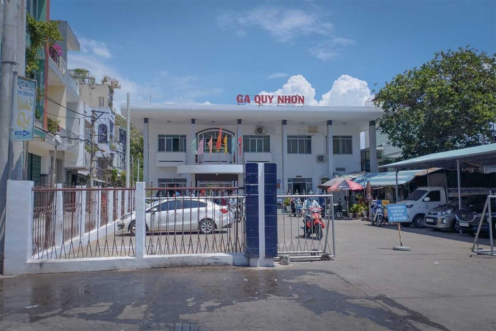 Exterior view of Quy Nhon Railway Station entrance on a sunny day, with motorbikes and cars parked outside the modest terminal.