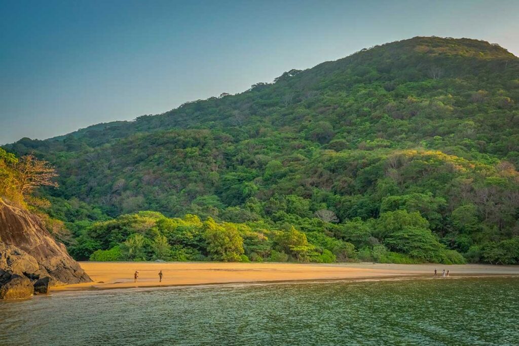 Remote Suoi Nong Beach in Con Dao with golden sand, lush forest-covered hills, and a few people walking along the quiet shoreline.