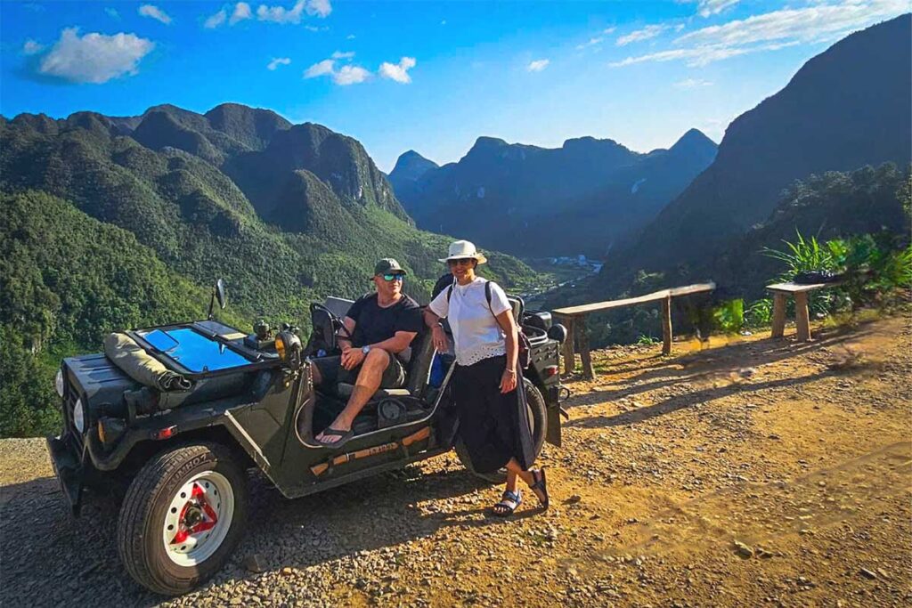 Two tourists posing near military jeep with Ha Giang mountain scenery in the background