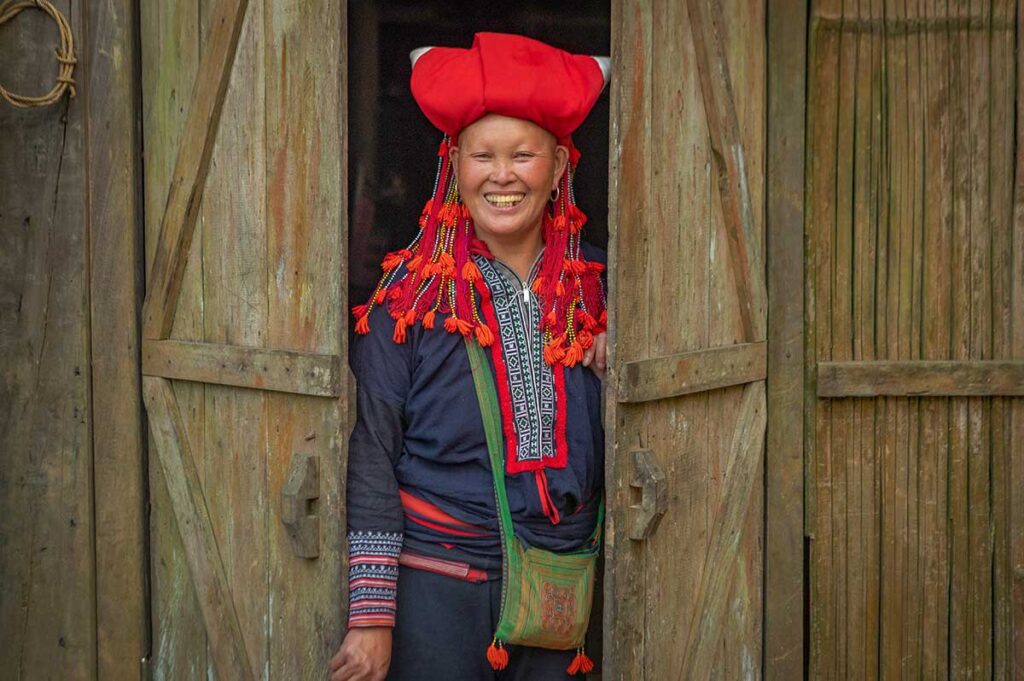 Smiling Red Dao woman standing in a wooden doorway in a rural Sapa village, featured on a Sapa homestay and trekking tour.