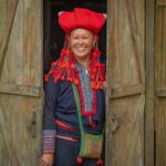 Smiling Red Dao woman standing in a wooden doorway in a rural Sapa village, featured on a Sapa homestay and trekking tour.